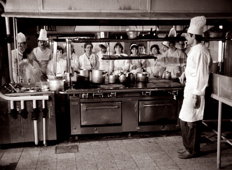 Grupa kucharzy i personelu kuchennego, wszyscy ubrani w mundury i czapki, stoją wokół dużego pieca w kuchni przemysłowej, podczas gdy jeden z kucharzy wydaje się instruować lub demonstrować coś grupie.