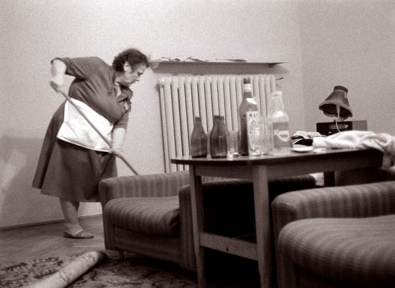 Starsza kobieta w sukience i fartuchu zamiata podłogę w salonie z fotelami, stołem pokrytym pustymi butelkami i grzejnikiem przy ścianie. Pokój wydaje się zagracony i słabo oświetlony.