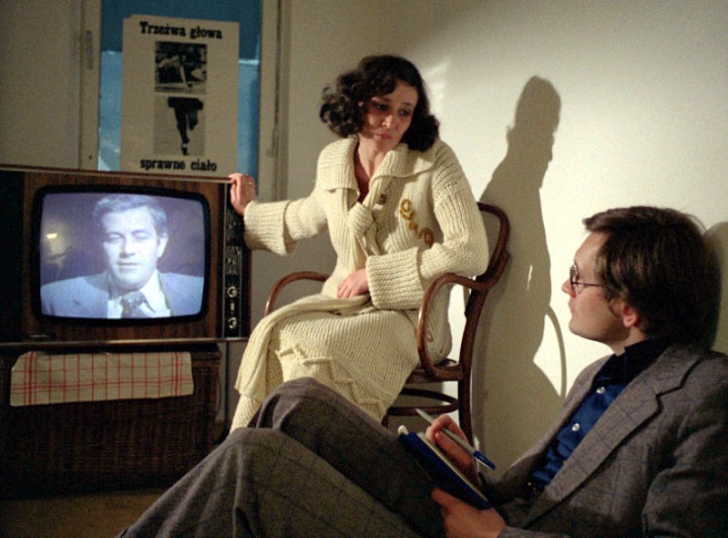 Kobieta w kremowym szlafroku siedzi na krześle obok telewizora, na którym widać twarz mężczyzny, podczas gdy mężczyzna w okularach siedzi na podłodze z notesem i patrzy na nią. Na ścianie za nimi wisi plakat z polskim tekstem.