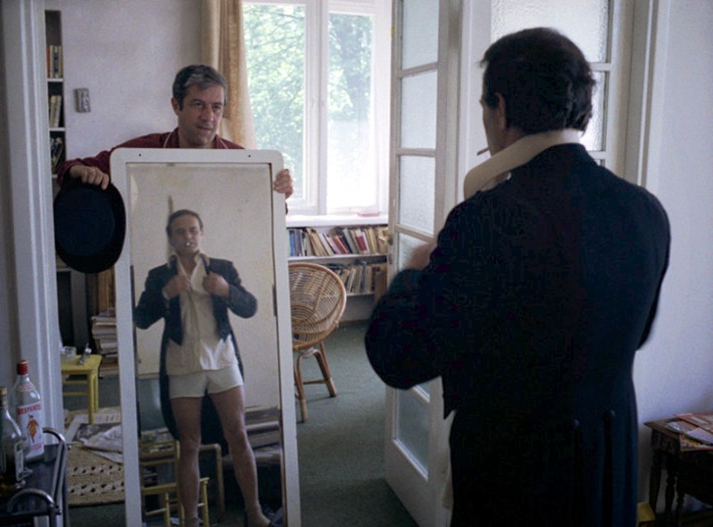 Mężczyzna trzyma lustro dla innego mężczyzny, który jest ubrany formalnie na górze, ale ma na sobie tylko bieliznę. Poprawia marynarkę w jasnym pokoju wypełnionym książkami.