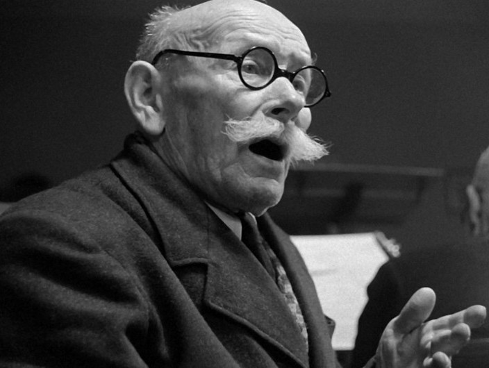 Starszy mężczyzna w okrągłych okularach i krzaczastym białym wąsie wygląda na zaskoczonego, z otwartymi ustami i podniesioną ręką, jakby mówił. Ma na sobie ciemny płaszcz i siedzi w pomieszczeniu przed rozmytym tłem.