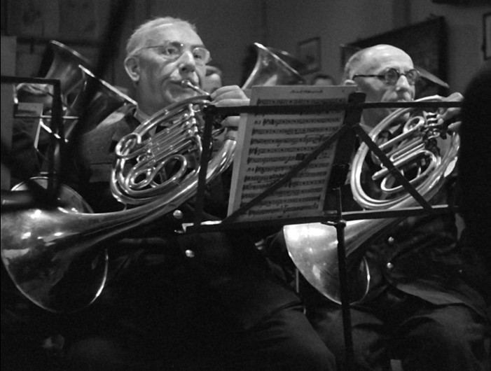 Dwóch starszych mężczyzn gra na waltorniach w orkiestrze. Obaj noszą okulary i ciemne garnitury, skupiając się na nutach umieszczonych na statywie przed nimi. Tło jest słabo oświetlone, a na ścianie widać rozmazane obrazy.