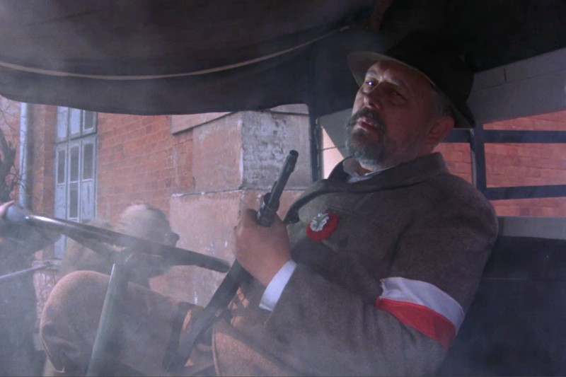 Starszy mężczyzna w kapeluszu i szarym płaszczu siedzi w zadymionym pojeździe, trzymając broń. Nosi czerwono-białą opaskę i przypinkę na klapie. W tle widać ceglane budynki.