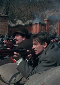 Dwóch mężczyzn w staromodnych płaszczach i kapeluszach kuca za workami z piaskiem, celując z karabinów podczas sceny bitwy rozgrywającej się na zewnątrz, w tle widać dym i zniszczone ceglane ściany.