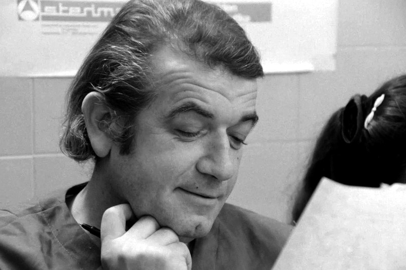 Mężczyzna o ciemnych włosach i bakach spogląda w dół na kartkę papieru, lekko się uśmiechając, z ręką opartą na brodzie. Nieopodal siedzi niewyraźna postać. Sceneria wydaje się być w pomieszczeniu, prawdopodobnie w biurze lub klasie.