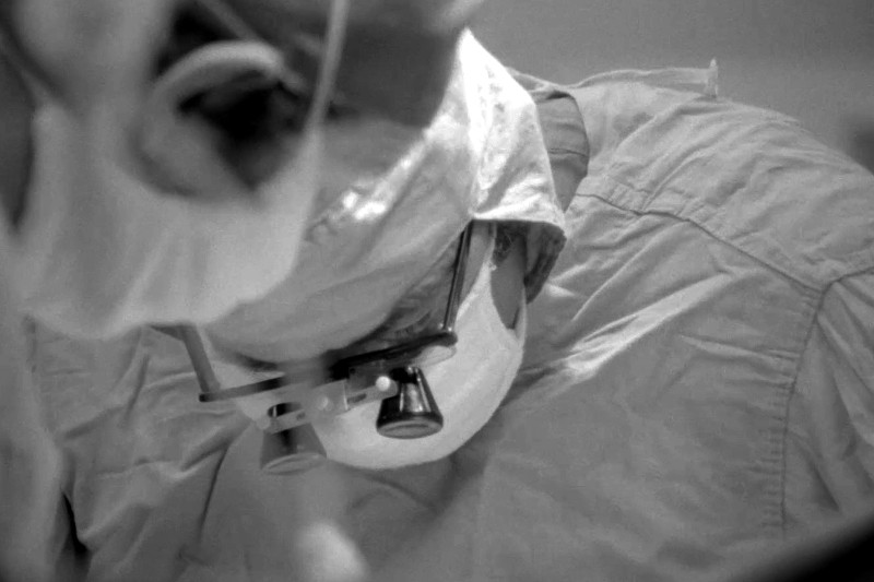 Chirurg w masce, czepku chirurgicznym i lupach skupia się intensywnie podczas zabiegu medycznego. Obraz jest czarno-biały.