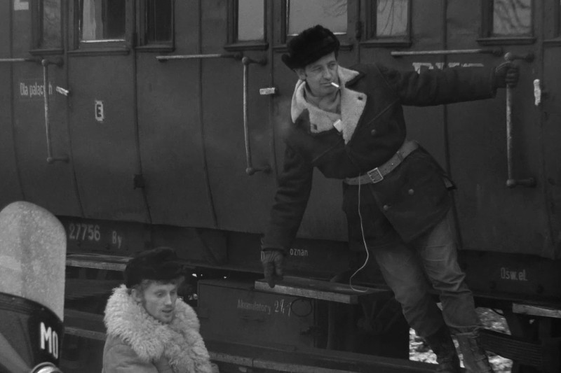 Dwóch mężczyzn w ciężkich płaszczach i futrzanych czapkach stoi przy pociągu; jeden pali papierosa i wychyla się ze schodów pociągu, podczas gdy drugi siedzi poniżej. Scena wydaje się rozgrywać w chłodne dni.