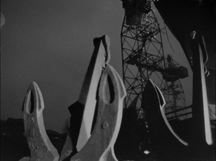 Duże kotwice statków na pierwszym planie są widoczne na tle ciemnego nieba, a w tle widać metalową wieżę radiową lub konstrukcję dźwigu. Obraz jest czarno-biały, co tworzy dramatyczny kontrast.