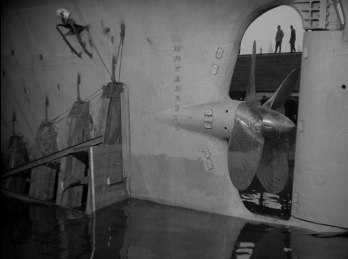 Zbliżenie na śrubę okrętową częściowo zanurzoną w wodzie, z widoczną częścią kadłuba. Dwie osoby stoją na nabrzeżu powyżej, a oznaczenia są widoczne na boku okrętu.