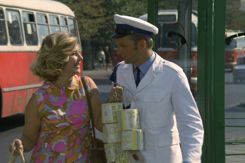 Kobieta w kolorowej, kwiecistej sukience uśmiecha się do mężczyzny w białym mundurze, trzymającego pliki pieniędzy i biletów, stojącego w pobliżu przystanku autobusowego, na tle którego widać czerwone i białe autobusy.