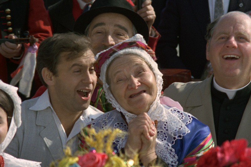 Grupa ludzi w odświętnych, tradycyjnych strojach uśmiecha się i śpiewa razem na zewnątrz, w środku znajduje się starsza kobieta w kolorowej chuście na głowie, radośnie ściskająca dłonie.