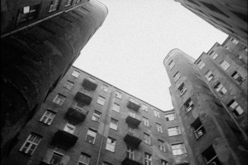Czarno-białe zdjęcie patrzące w górę na wysokie, stare ceglane budynki mieszkalne tworzące dziedziniec. Budynki mają wiele okien i balkonów, a niebo nad nimi jest zachmurzone.