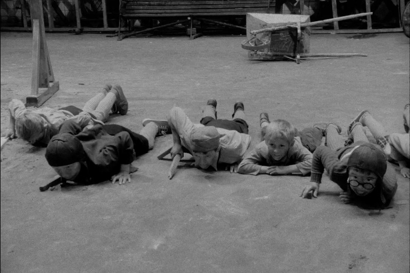 Pięcioro dzieci leży na brzuchu na betonowej podłodze, zwróconych twarzą do przodu. Wyglądają, jakby raczkowały lub się bawiły, i są ubrane w codzienne ubrania typowe dla połowy XX wieku. W tle widać ławkę i drewniane skrzynki.