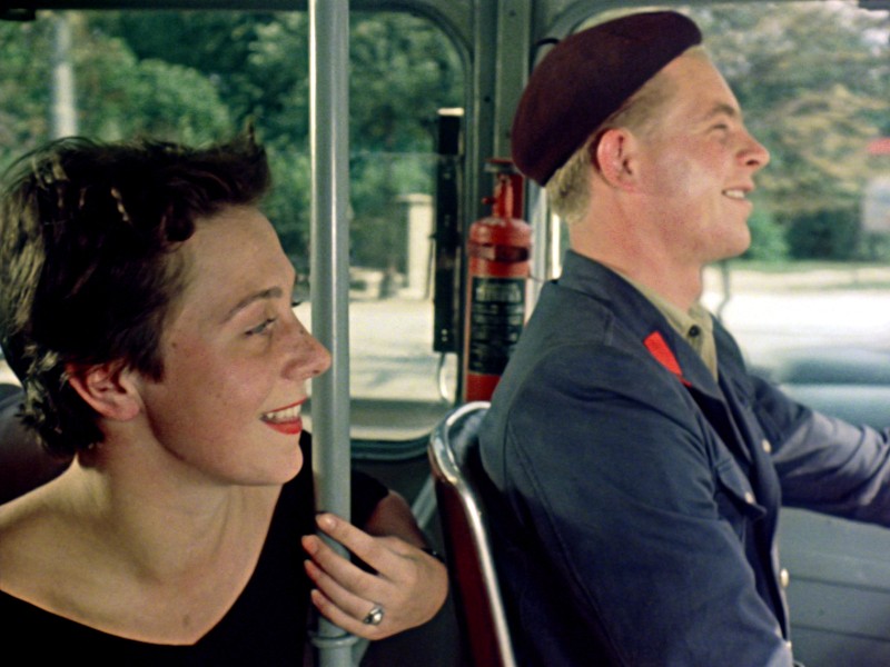Kobieta z krótkimi brązowymi włosami uśmiecha się do mężczyzny w mundurze i berecie, który prowadzi pojazd. Scena wydaje się radosna, a światło słoneczne sączy się przez okna autobusu, a na zewnątrz widać zielone drzewa.