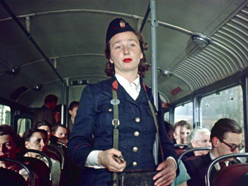 Kobieta-konduktorka w granatowym mundurze stoi w przejściu zatłoczonego autobusu, trzymając w ręku kasownik biletu, a pasażerowie siedzą w rzędach za nią.