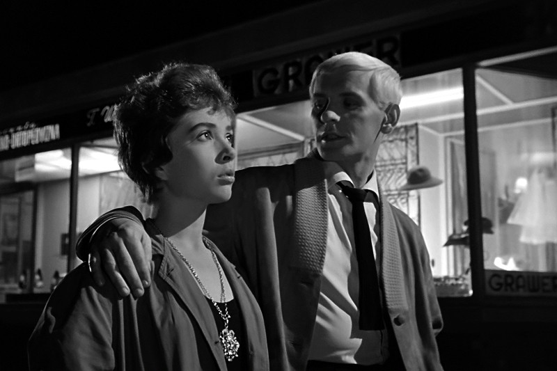 Mężczyzna o jasnych włosach obejmuje kobietę, stojąc nocą przed jasno oświetlonym sklepem. Kobieta patrzy poważnie przed siebie, podczas gdy mężczyzna do niej mówi.