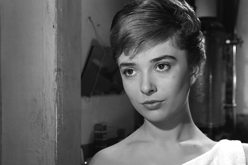 Młoda kobieta z krótkimi, starannie ułożonymi włosami patrzy lekko na bok, ma na sobie biały ręcznik przerzucony przez ramiona. W tle znajdują się przedmioty gospodarstwa domowego w delikatnie oświetlonym, czarno-białym otoczeniu.