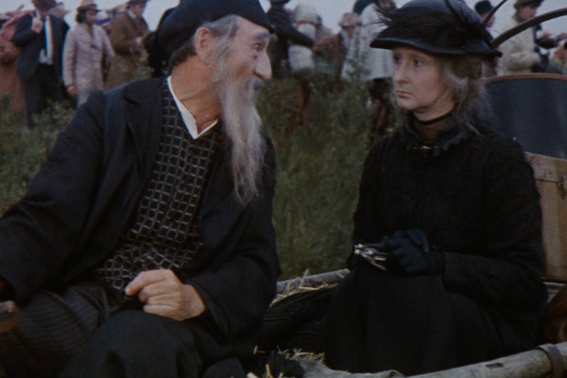 Starszy mężczyzna z długą białą brodą rozmawia ożywiony z kobietą ubraną na czarno, w kapeluszu i rękawiczkach, która patrzy na niego poważnie. Siedzą na zewnątrz w tłumie, prawdopodobnie na jakimś wydarzeniu lub spotkaniu.