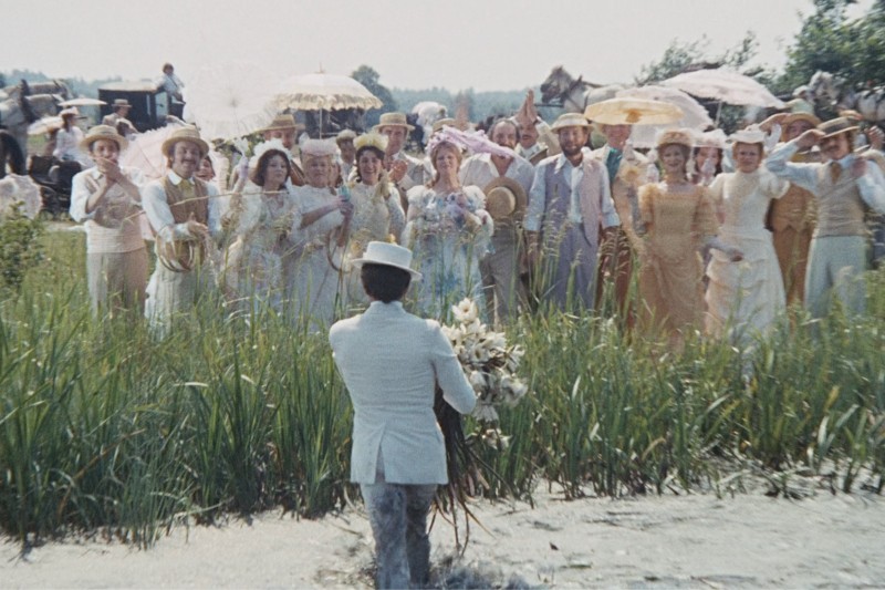 Mężczyzna w jasnym garniturze trzymający kwiaty zbliża się do grupy ludzi ubranych w eleganckie pastelowe ubrania, niektórzy z nich trzymają parasole, stojących na trawiastym polu w słoneczny dzień.