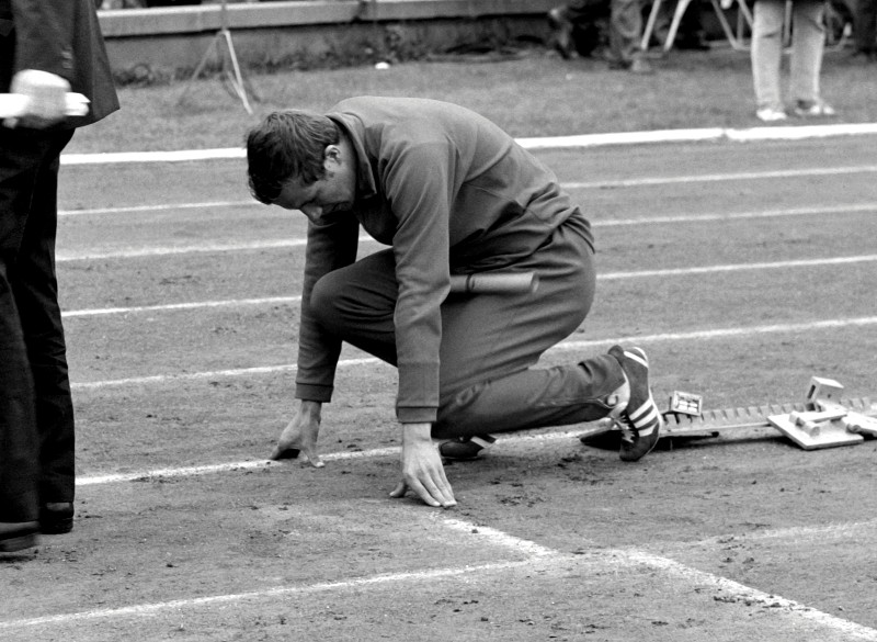 Lekkoatleta w dresie klęka na blokach startowych na bieżni, przygotowując się do wyścigu. Scena jest czarno-biała, a sportowiec wydaje się skupiony i gotowy.