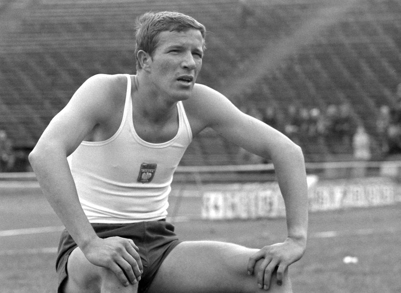 Sportowiec w podkoszulku i krótkich spodenkach odpoczywa z jednym kolanem na bieżni, patrząc przed siebie ze skupionym wyrazem twarzy. W tle widać puste siedzenia stadionu i kilka osób w oddali. Obraz jest czarno-biały.