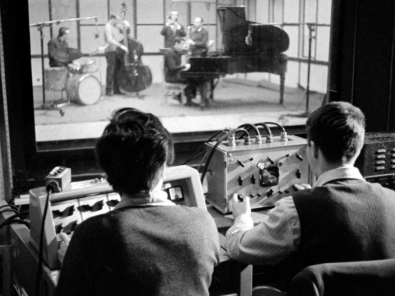 Dwie osoby obsługują sprzęt audio w pokoju kontrolnym, jednocześnie oglądając przez duże okno występ zespołu jazzowego na żywo, składającego się z pianisty, basisty, perkusisty i muzyków dętych, występującego w studiu.