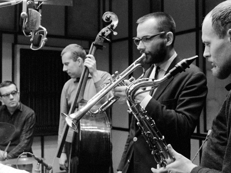 Czterech muzyków występuje w studiu; jeden gra na perkusji, drugi na kontrabasie, a dwóch na instrumentach dętych, w tym na trąbce i saksofonie. Widoczne są mikrofony i panele dźwiękoszczelne.