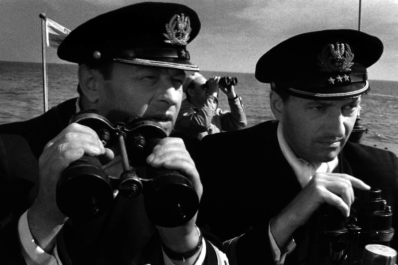 Dwóch mężczyzn w mundurach marynarki wojennej patrzy przez lornetkę na statek, w tle widać ocean i innych członków załogi. Obraz jest czarno-biały.