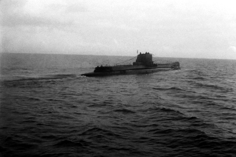 Czarno-białe zdjęcie częściowo wynurzonej łodzi podwodnej poruszającej się po spokojnych, otwartych wodach oceanu pod zachmurzonym niebem.