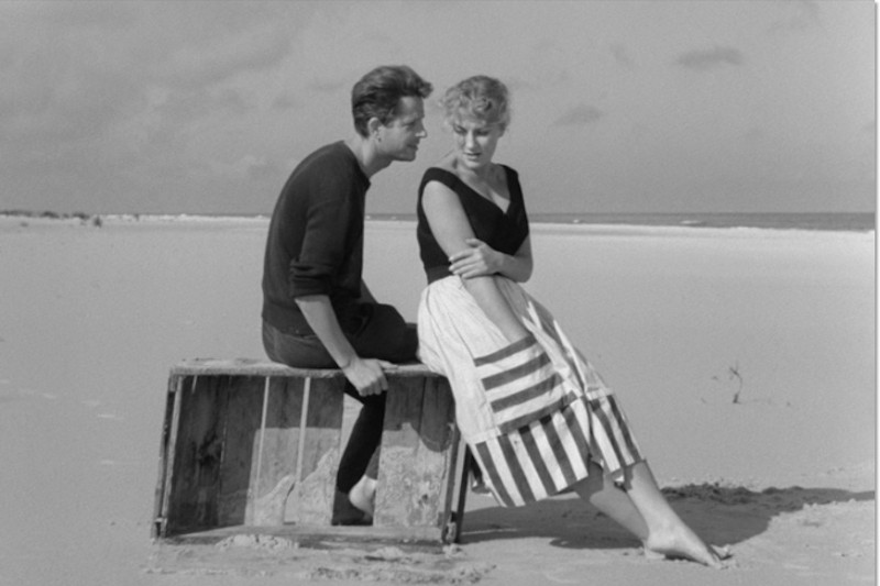 Mężczyzna i kobieta siedzą na drewnianej skrzyni na pustej piaszczystej plaży. Mężczyzna pochyla się w stronę kobiety, a ona zamyślona odwraca wzrok. Oboje są boso i ubrani w ubrania w stylu lat 50. Niebo jest częściowo zachmurzone.