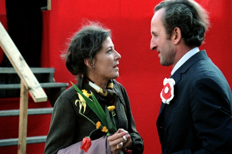 Kobieta trzymająca tulipany i żonkile rozmawia z mężczyzną w garniturze z biało-czerwoną rozetą na klapie. Stoją przed jaskrawo czerwonym tłem z drewnianymi schodami po lewej stronie.