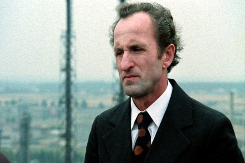 Mężczyzna w średnim wieku w ciemnym garniturze i wzorzystym krawacie stoi na zewnątrz, na tle zachmurzonego nieba widać rozmyte wieże przemysłowe. Ma krótkie, kręcone włosy i poważny wyraz twarzy.