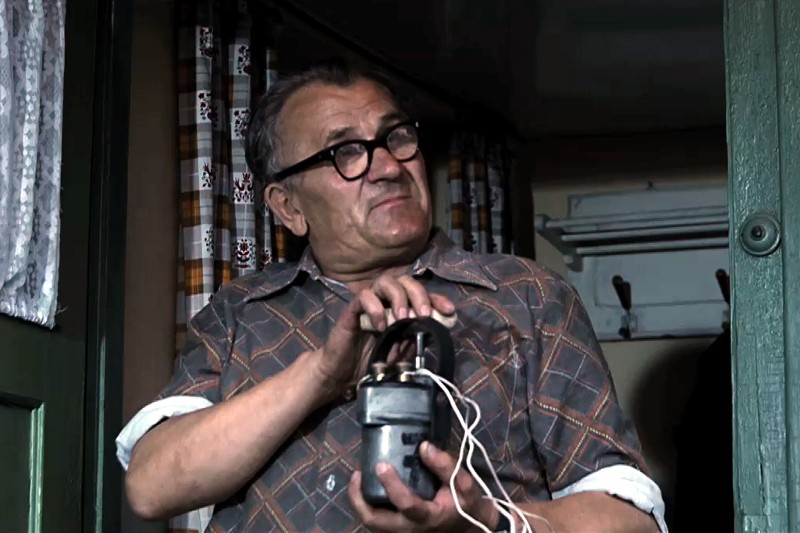 Starszy mężczyzna w okularach i kraciastej koszuli stoi w pomieszczeniu, trzymając w dłoniach niewielkie urządzenie z przymocowanymi przewodami, przy zielonych drzwiach i wzorzystych zasłonach w tle.