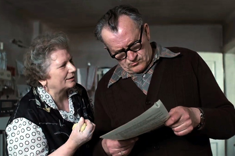 Starsza kobieta uśmiecha się i gestykuluje lewą ręką, rozmawiając ze starszym mężczyzną w okularach, który jest skupiony na czytaniu dokumentu, który trzyma. Stoją w pomieszczeniu, w skromnie umeblowanym pokoju.