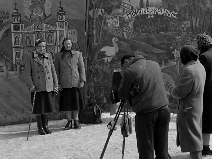 Dwie kobiety pozują do zdjęcia na tle malowanego tła, podczas gdy fotograf robi im zdjęcie na zewnątrz. Dwie inne osoby stoją w pobliżu i obserwują. Ziemię pokrywa śnieg. W tle widać budynki i napis Pamiątka z Kalwarii 1952.