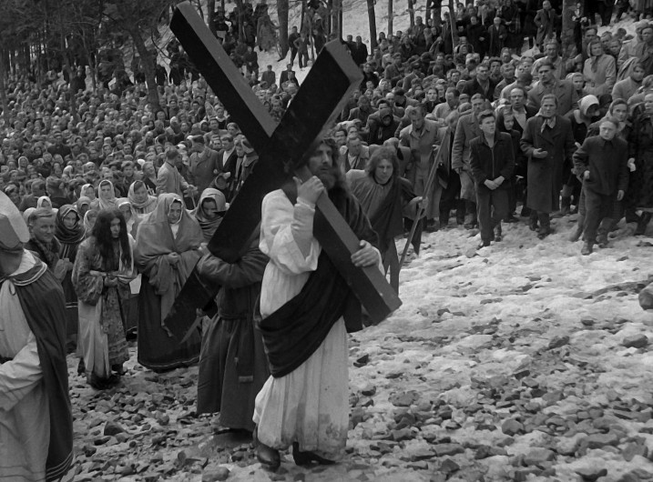 Mężczyzna ubrany w strój biblijny niesie duży drewniany krzyż na ośnieżone, skaliste wzgórze, za nim podąża grupa ludzi, a w tle obserwuje go duży tłum ludzi.