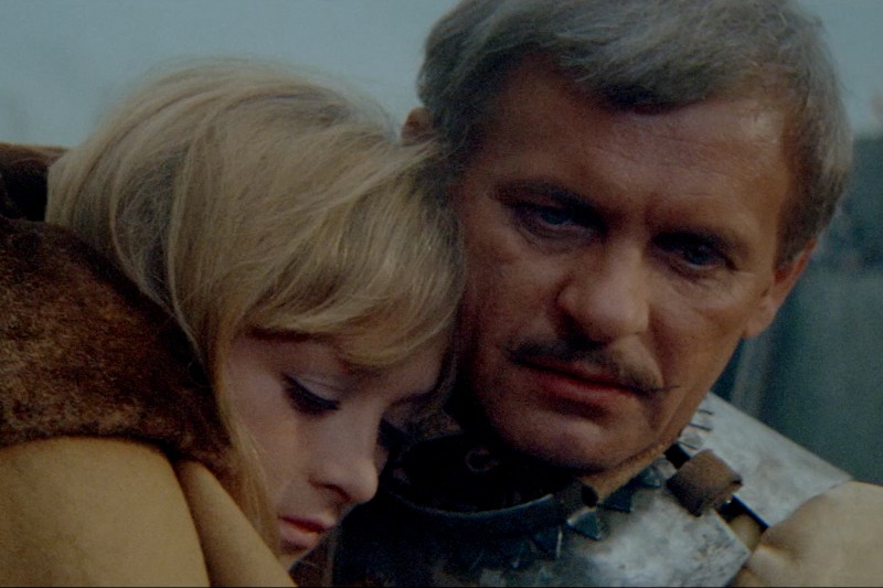 Mężczyzna w średniowiecznej zbroi z wąsami wygląda poważnie, obejmując kobietę o blond włosach, która opiera głowę na jego ramieniu. Oboje wydają się emocjonalni i blisko siebie.