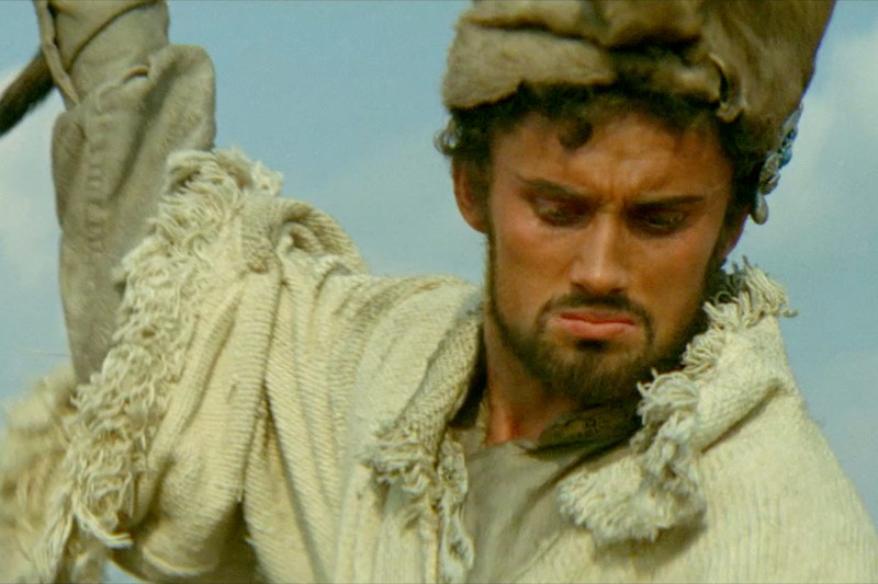 Mężczyzna z kręconymi ciemnymi włosami i brodą nosi futrzaną czapkę i szorstkie, frędzlowane, kremowe ubranie. Patrzy w dół z poważnym wyrazem twarzy, jakby w ruchu. W tle widać błękitne niebo.