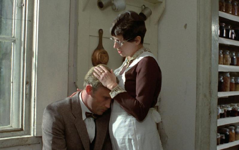 Mężczyzna w garniturze siedzi z głową opartą o klatkę piersiową kobiety. Kobieta w okularach i ciemnej sukience z białym fartuchem delikatnie trzyma jego głowę. Znajdują się w rustykalnej kuchni z półkami słoików i wiszącymi kubkami.