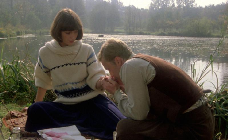 Kobieta w białym swetrze siedzi nad jeziorem, a mężczyzna obok niej klęka i całuje ją w rękę. Robią piknik na trawie, a w tle widać drzewa, wodę i grzybienie.