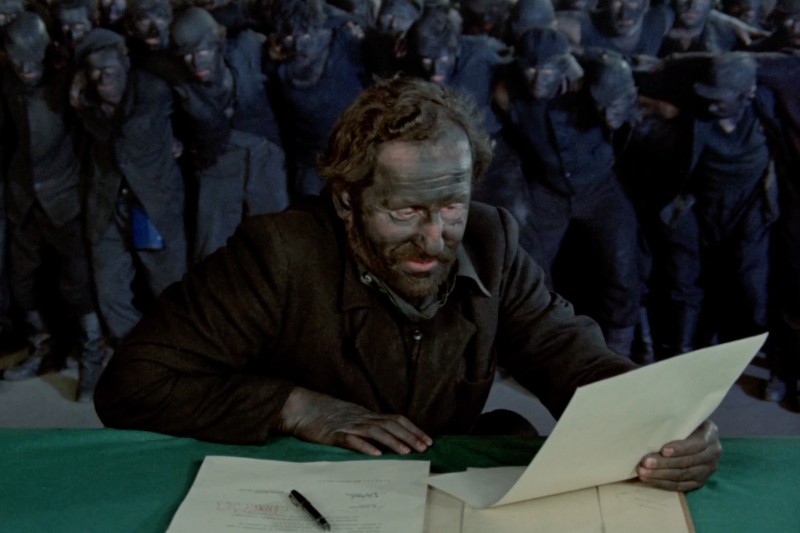 Osoba z twarzą i dłońmi pokrytymi czarną sadzą czyta dokument przy biurku; za nimi grupa podobnie pokrytych sadzą osób stoi blisko siebie, obserwując. Na biurku leżą papiery i długopis.