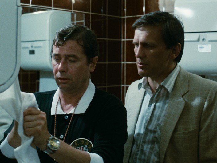 Dwóch mężczyzn stoi w łazience z brązowymi kafelkami na ścianach i białymi suszarkami do rąk. Jeden mężczyzna wyciera ręce ręcznikiem papierowym, podczas gdy drugi stoi tuż obok niego, uważnie mu się przyglądając.