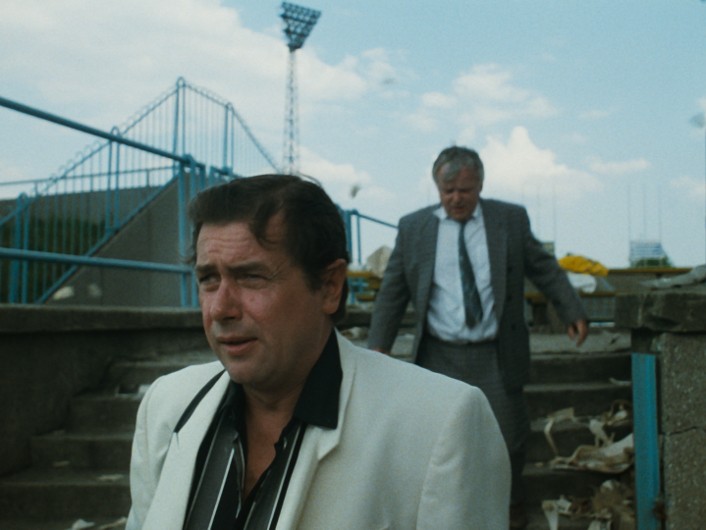 Dwóch mężczyzn w garniturach schodzi po schodach stadionu na zewnątrz, jeden w białej kurtce na pierwszym planie wygląda na zaniepokojonego, drugi w szarym garniturze podąża za nim. Niebo jest czyste, nad nim jest kilka chmur.