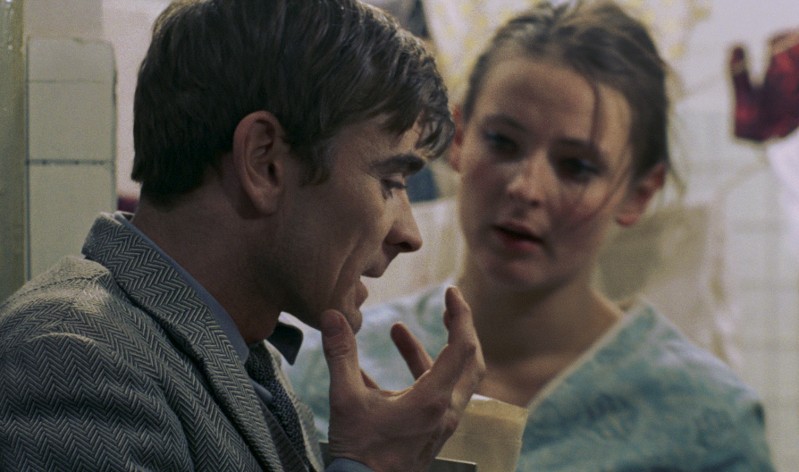 Mężczyzna we wzorzystym garniturze dotyka swojej twarzy, rozmawiając z kobietą w niebieskiej bluzce, która patrzy na niego z niepokojem w łagodnie oświetlonym wnętrzu.