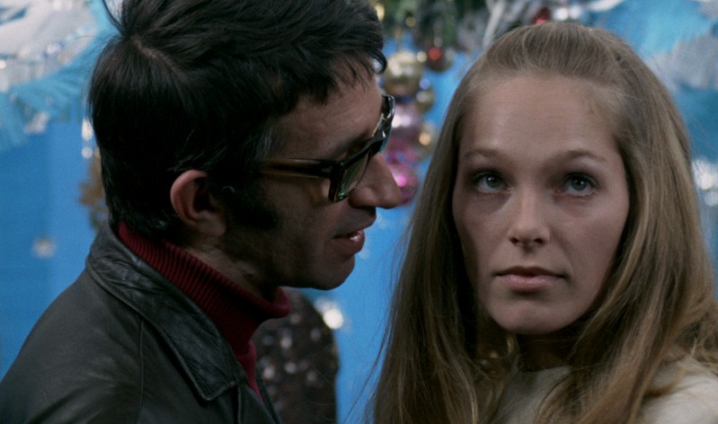 Mężczyzna w okularach i ciemnej kurtce pochyla się w stronę kobiety o długich blond włosach, która patrzy prosto przed siebie. Stoją przed niebieskim tłem z udekorowaną choinką.