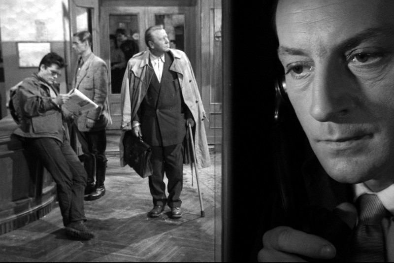 Mężczyzna trzyma słuchawkę telefonu w kabinie po prawej stronie, podczas gdy trzej mężczyźni stoją i rozmawiają przy biurku w holu po lewej stronie. Wszystko to w czarno-białym otoczeniu.