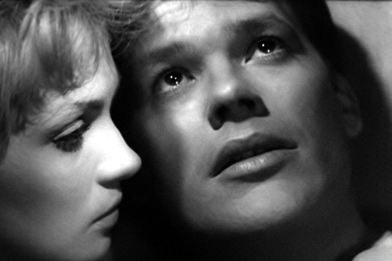 Czarno-białe zdjęcie przedstawiające dwie osoby z bliska; kobieta po lewej uważnie wpatruje się w mężczyznę po prawej, którego twarz jest oświetlona, a on sam patrzy w górę z emocjonalnym, refleksyjnym wyrazem twarzy.