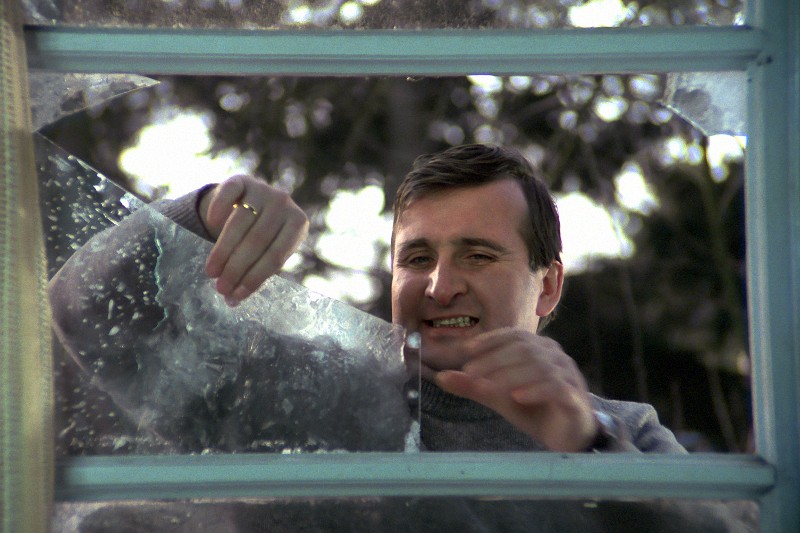 Mężczyzna krzywi się, usuwając duży, nierówny kawałek stłuczonego szkła z szyby okiennej. W tle widać drzewa.