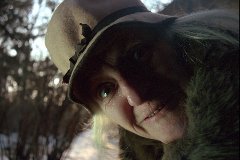 Osoba z długimi, jasnymi włosami nosi kapelusz z szerokim rondem i płaszcz z futrzanym kołnierzem, lekko się uśmiecha, patrząc uważnie w obiektyw. W tle widać drzewa i śnieżną scenerię na zewnątrz.