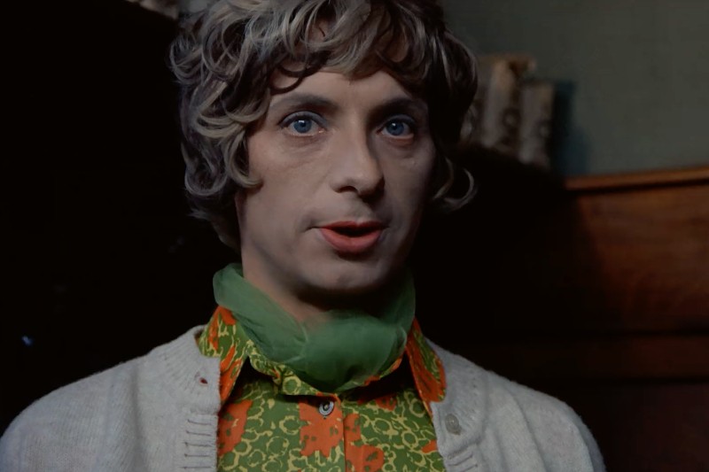 Osoba o kręconych, jasnobrązowych włosach i niebieskich oczach, ubrana w zielony szalik, wzorzystą pomarańczowo-zieloną koszulę i beżowy sweter, z delikatnym makijażem i jasnoczerwoną szminką, stoi w pomieszczeniu.